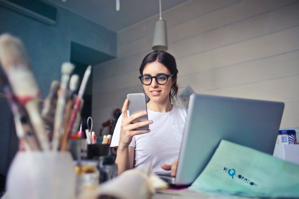 bruce-mars-FWVMhUa_wbY-unsplash female in front of laptop with pens and brushes in the foreground. Female is holding a smartphone.