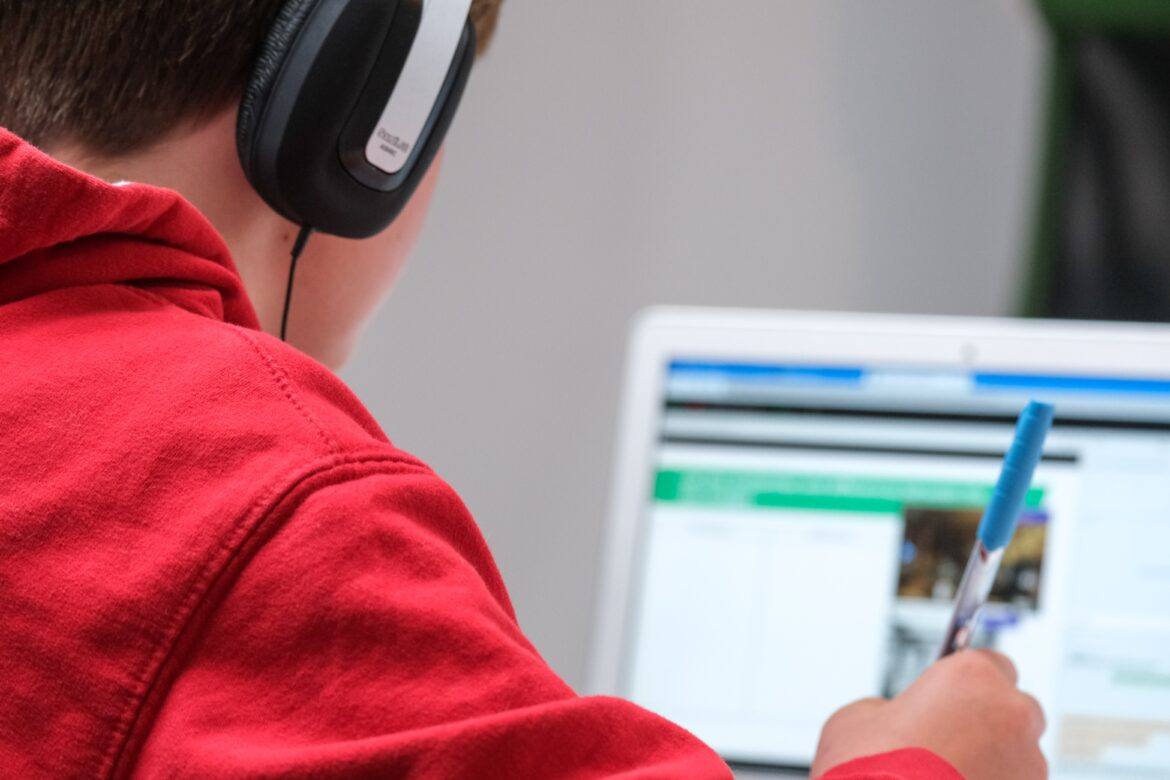 compare-fibre-Y8TiLvKnLeg-unsplash Student with headphones, red jacket, holding a pen, while looking at a laptop screen