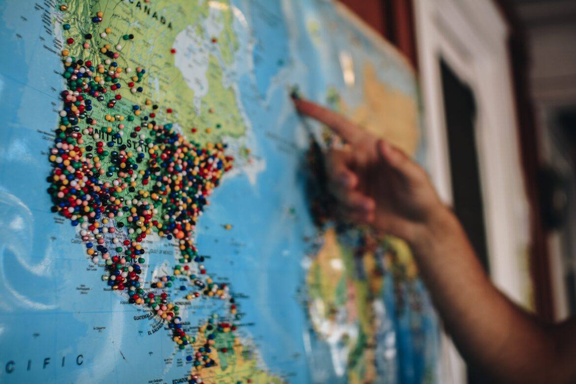 kelsey-knight-SFRw5GChoLA-unsplash World map with pushpins through-out. a human hand with finger pointing to a pushpin appears.