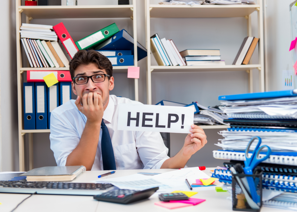 Stressed male employee at a messy desk with piles of work surrounding him holds up a sign that says, "HELP!"