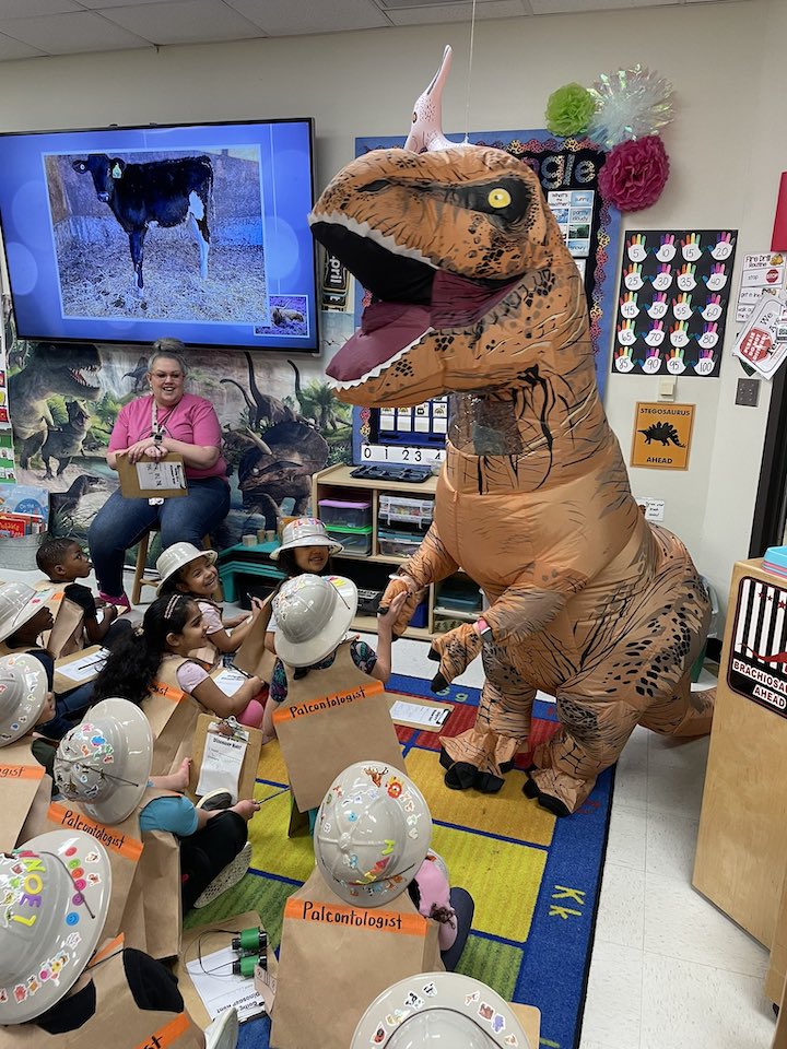 Students in class wearing safari attire with a teacher and an adult in a blow up dinosaur costume.