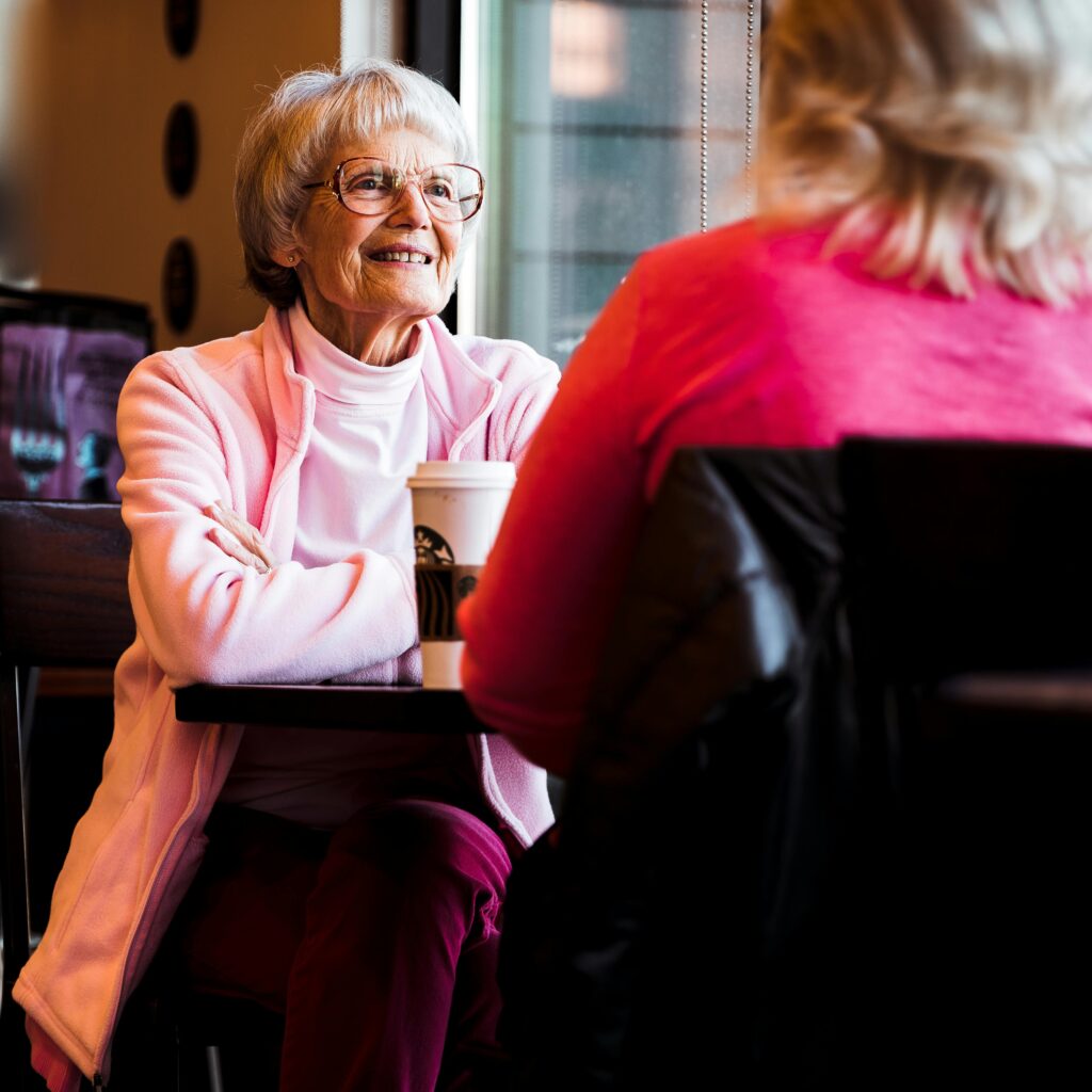 An elderly woman wearing glasses and a pink jacket smiles while sitting at a table in a café, holding a coffee cup, across from another person