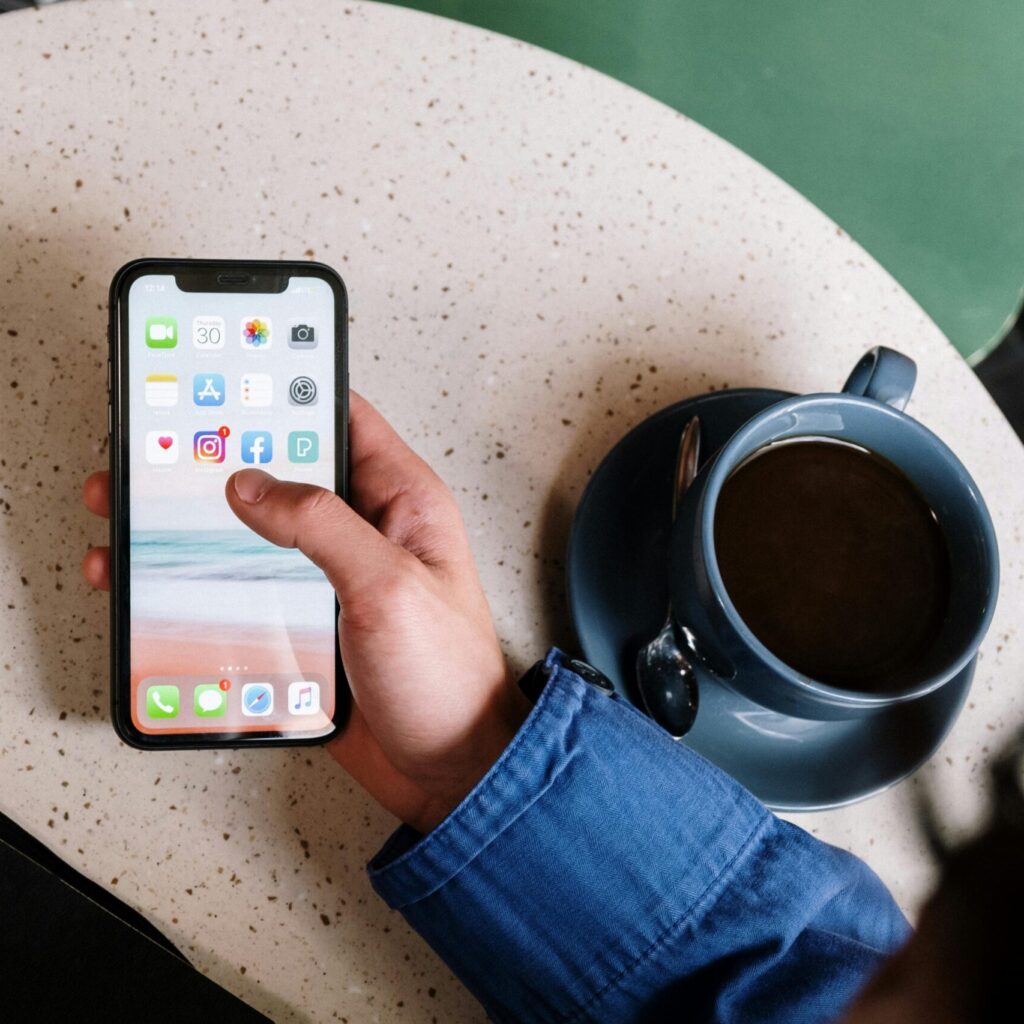 A person in a blue jacket holds a smartphone displaying app icons, with a cup of coffee, a notebook, and a pen on a round table