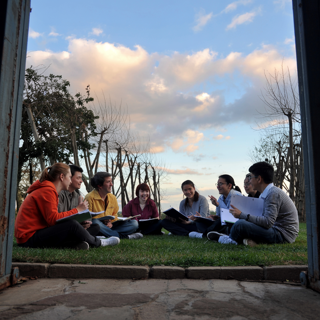 A group of students sits in a circle on a grassy lawn, holding notebooks and engaging in lively discussion under a sky with soft clouds and bare trees in the background.