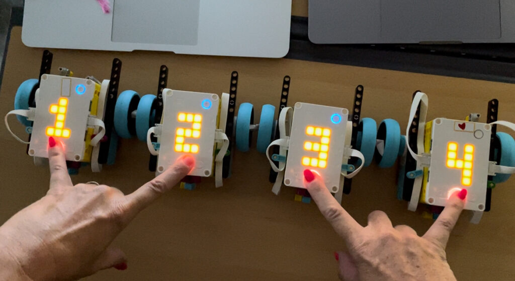 Four small wheeled robots lined up on a table, each displaying a bright LED number from 1 to 4. Hands with red-painted nails are pressing buttons on each robot's faceplate.