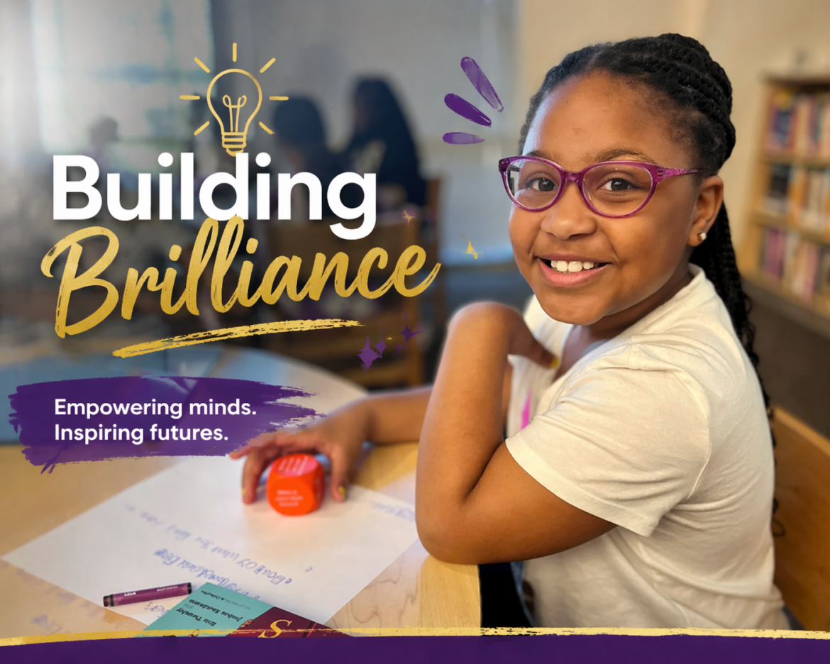 Smiling elementary student wearing purple glasses works at a desk in a classroom with the text “Building Brilliance” overlaid on the image.