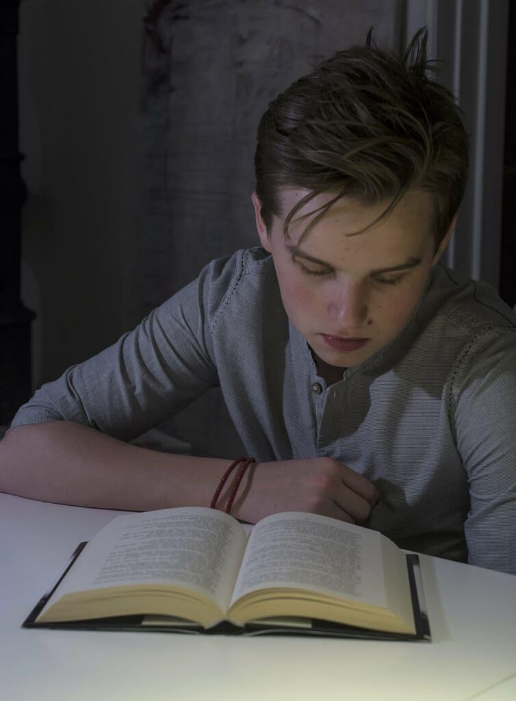 Young person with dark hair leans over an open book at a white table, reading intently in a softly lit, shadowed room.